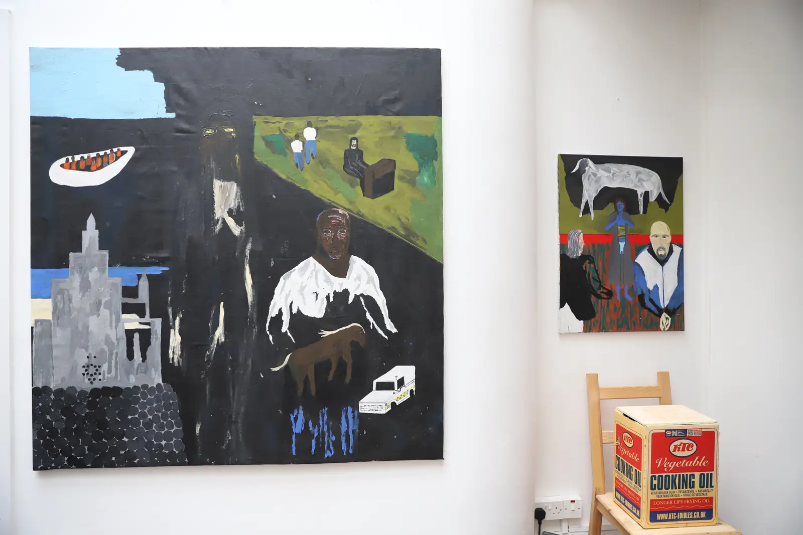 Installation view of Paes’ paintings displayed with everyday objects, including a chair and cardboard box, inside a gallery.