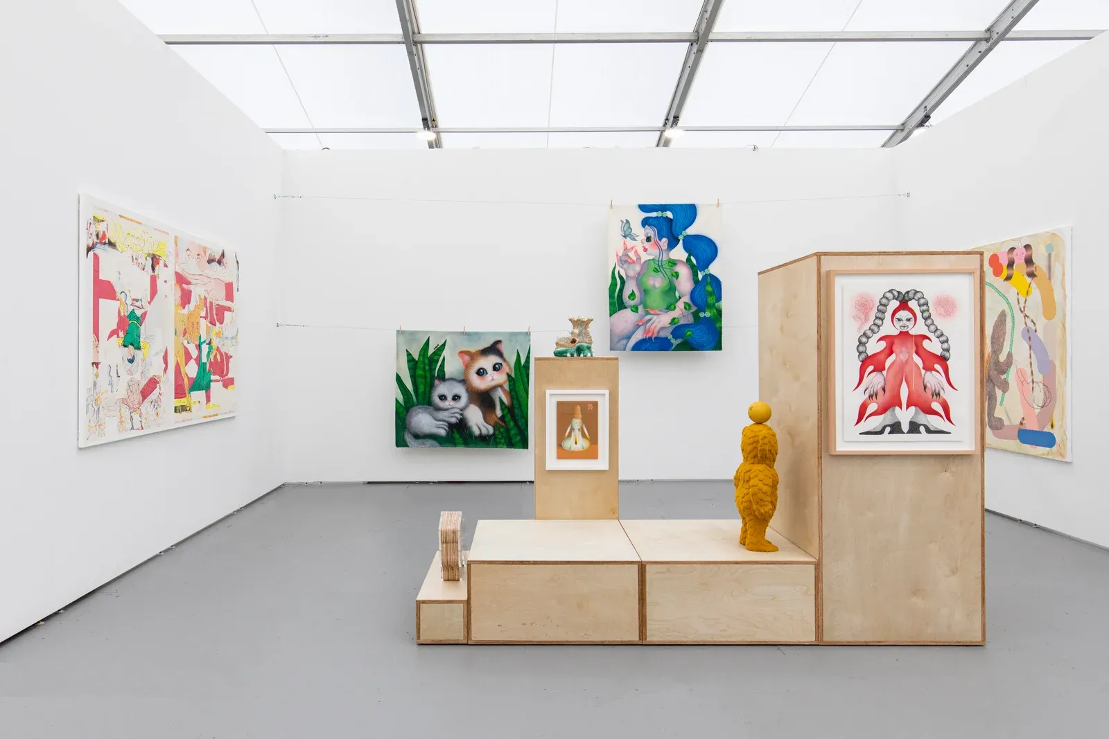 Installation view of works by Fátima de Juan presented on wooden plinths and walls, combining figurative paintings and sculptural elements in a bright, booth-like exhibition setting.