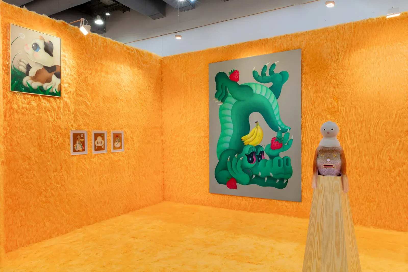 Detail view of Fátima de Juan’s presentation at Zona Maco Art Fair, Mexico, showing a green creature-like painting with fruit motifs alongside sculptural figures, installed against textured orange walls.