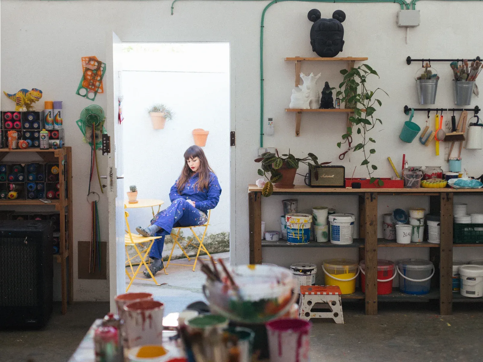 Fátima de Juan, Portrait of the artist seated in the doorway of her studio, surrounded by shelves, paint buckets, and studio tools.