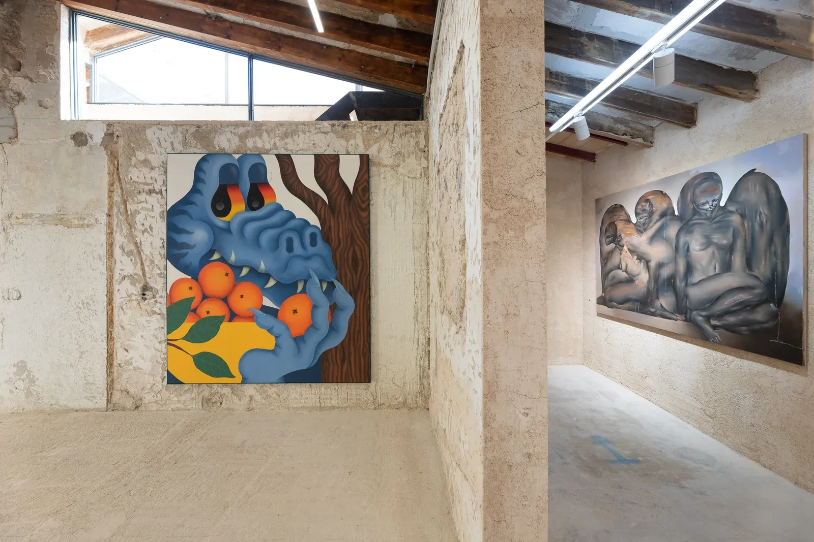 Installation view of Fátima de Juan’s La cosecha, a large-scale acrylic and spray painting depicting a blue, creature-like figure holding oranges, shown on a textured gallery wall within an industrial exhibition space.
