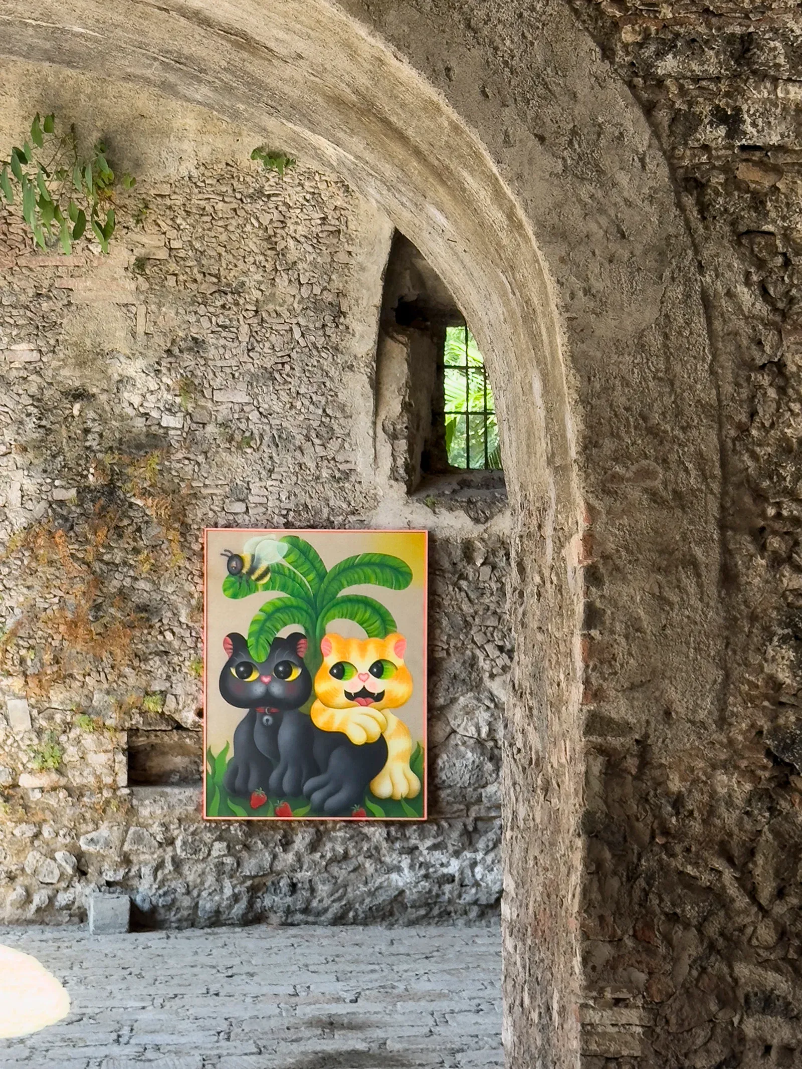 Installation view of Fátima de Juan’s painting showing two cat-like figures beneath palm leaves, installed within the stone interior of Hacienda Acamilpa, combining figurative imagery with historic architecture.
