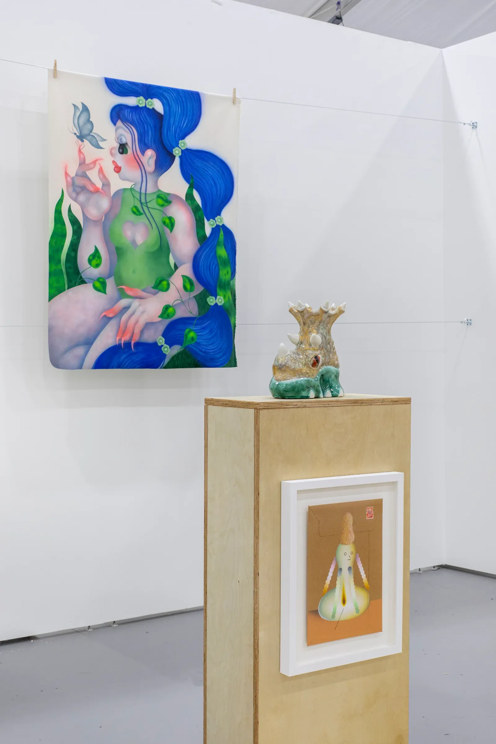 Installation view featuring a figurative painting of a blue-haired female figure alongside small sculptural works displayed on wooden plinths, highlighting the dialogue between painting and sculpture.