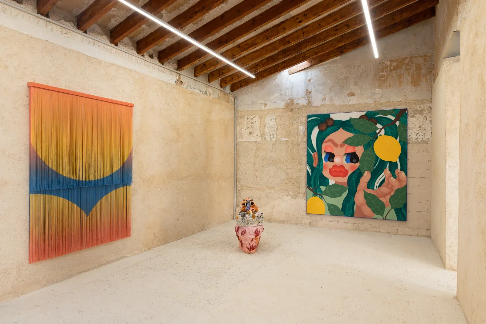 Installation view of Fátima de Juan’s work in the exhibition Entre Cajas, showing a large figurative painting with fruit motifs alongside a ceramic sculpture placed in a raw, vaulted interior space.