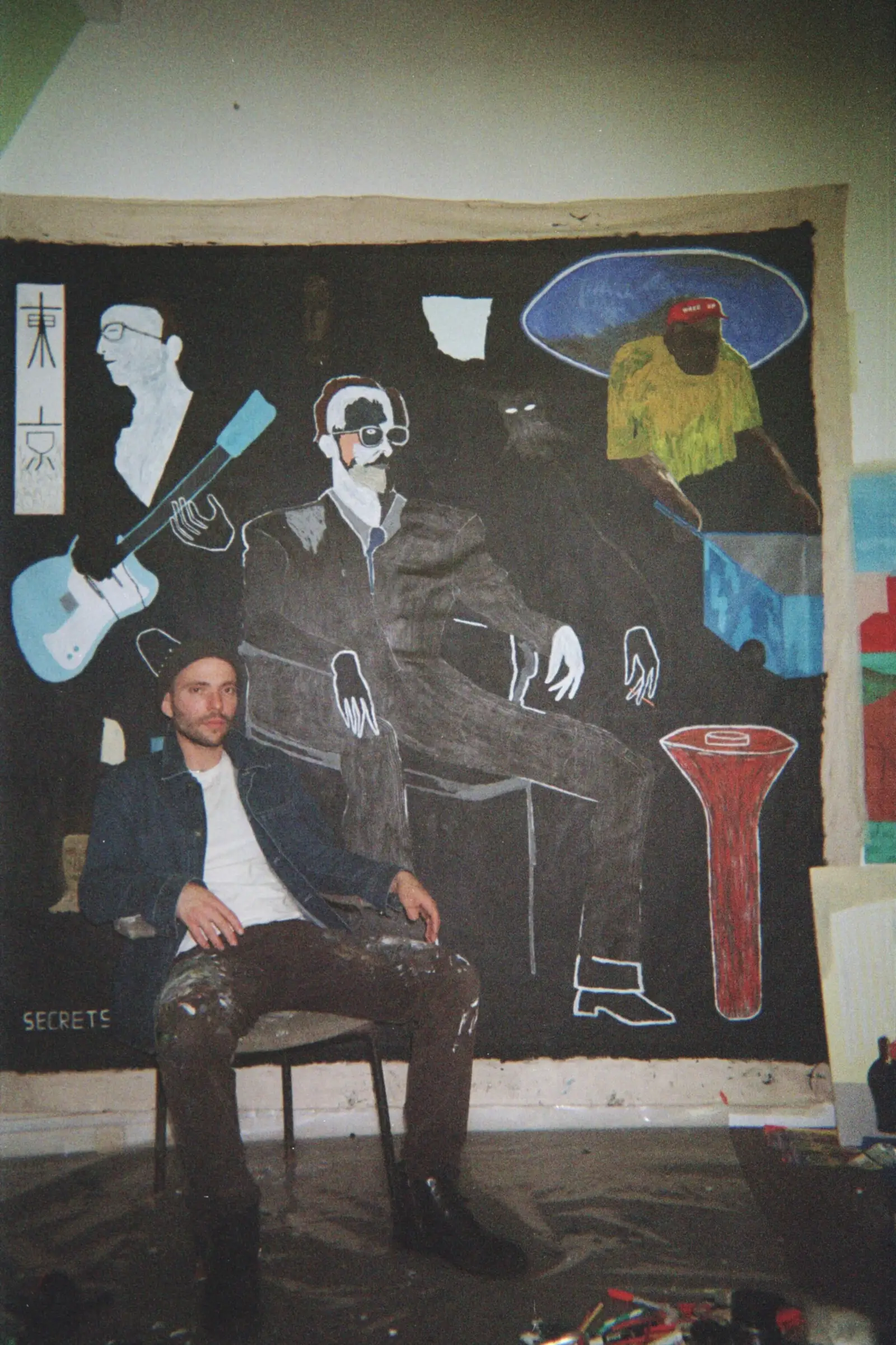 contemporary artist Paes sitting on a chair infront of his artwork Secrets of Unfolding Leisure