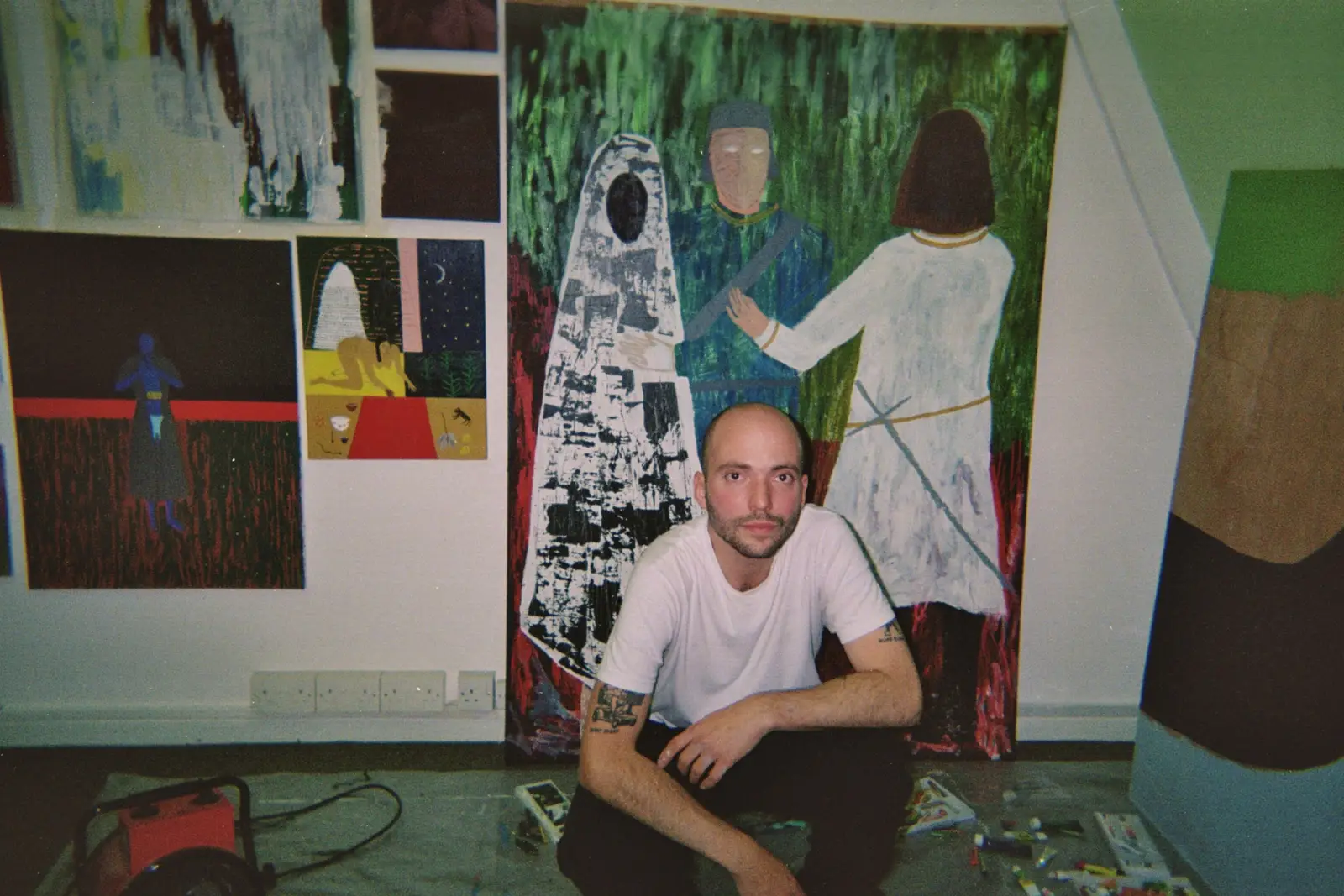 Portrait of artist Paes seated on a chair in front of a large black painting depicting a seated figure and abstract characters, photographed in the studio.