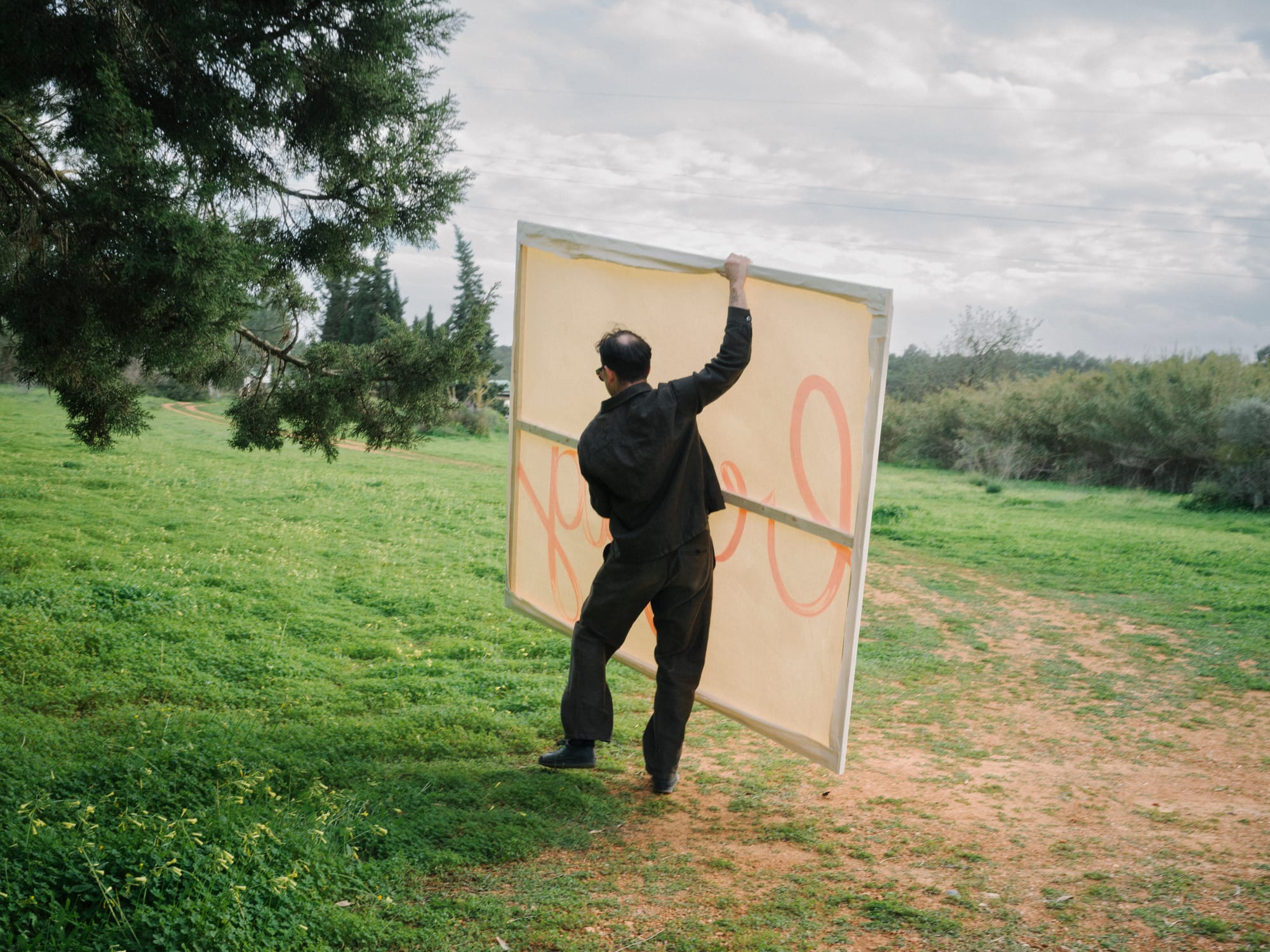 Artist Tom Król outdoors holding a large canvas with orange lettering visible on the surface, standing in a grassy landscape under a cloudy sky