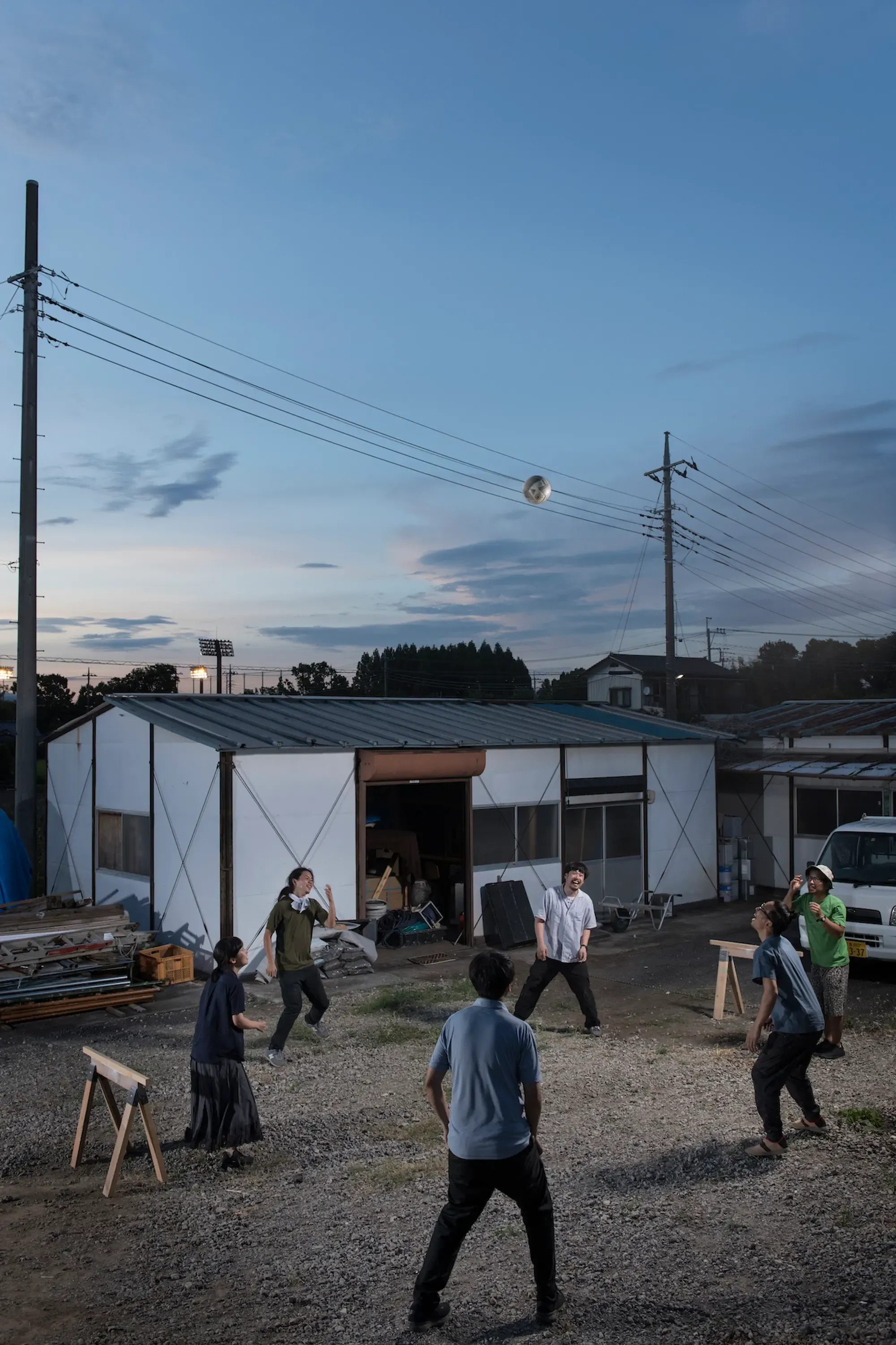 目 [mé] and collaborators playing outdoors at dusk in front of their studio building, a reflective sphere suspended in the sky above an industrial courtyard.