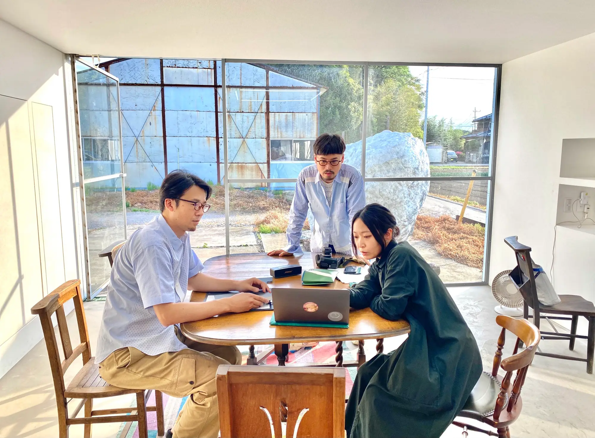 目 [mé] team gathered around a wooden table inside a glass-walled workspace, reviewing material on a laptop with sculptural forms visible outside.