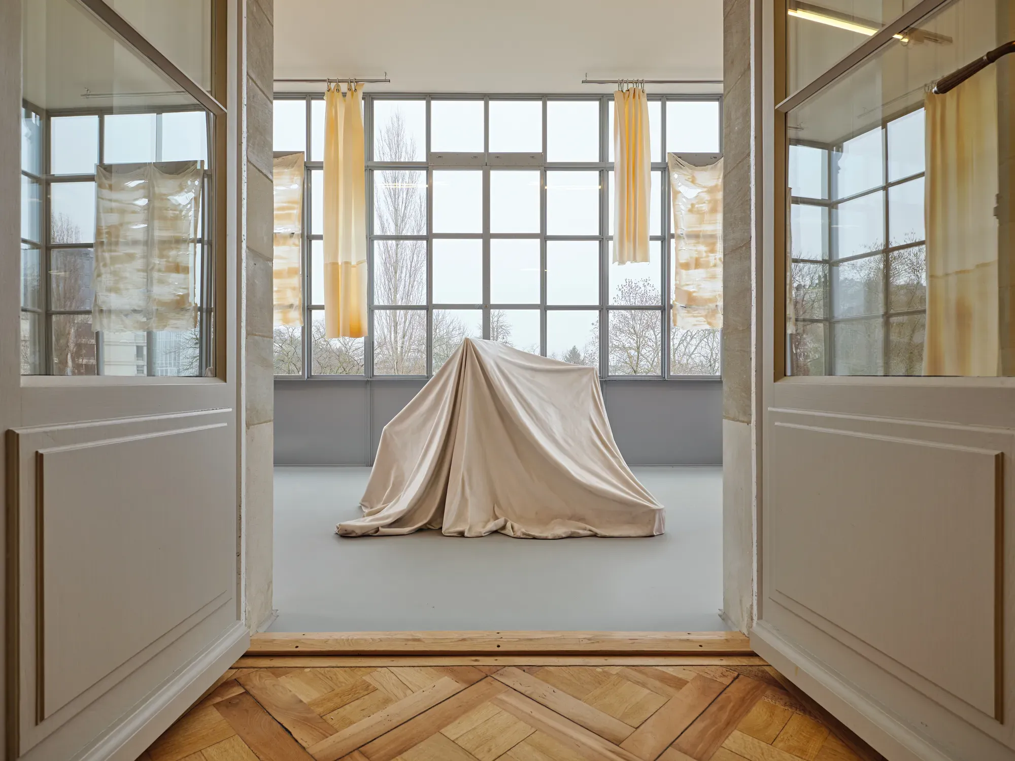 Ryan Gander sculpture covered with fabric placed in the center of a gallery room at Villa Bernasconi, surrounded by Li Tavor’s translucent latex curtain works hanging in front of large windows in the exhibition “Stitches: A Haunted House and Other Ghost Stories”, 2026.