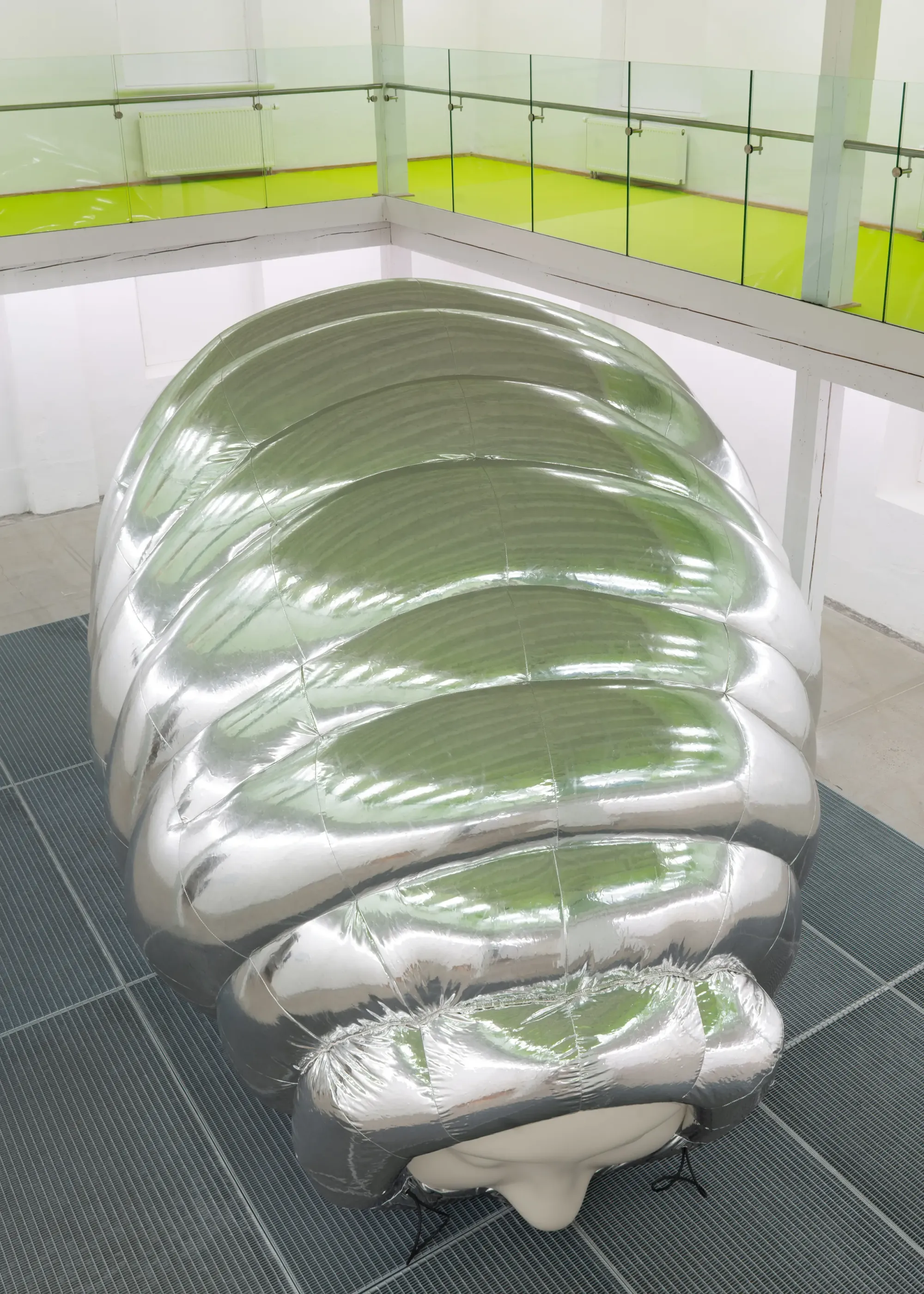 Top view of large inflatable silver cocoon sculpture with segmented reflective surface and partially visible human face, placed on dark floor grid in gallery setting, Stine Deja Micro Management Viborg Kunsthal Denmark
