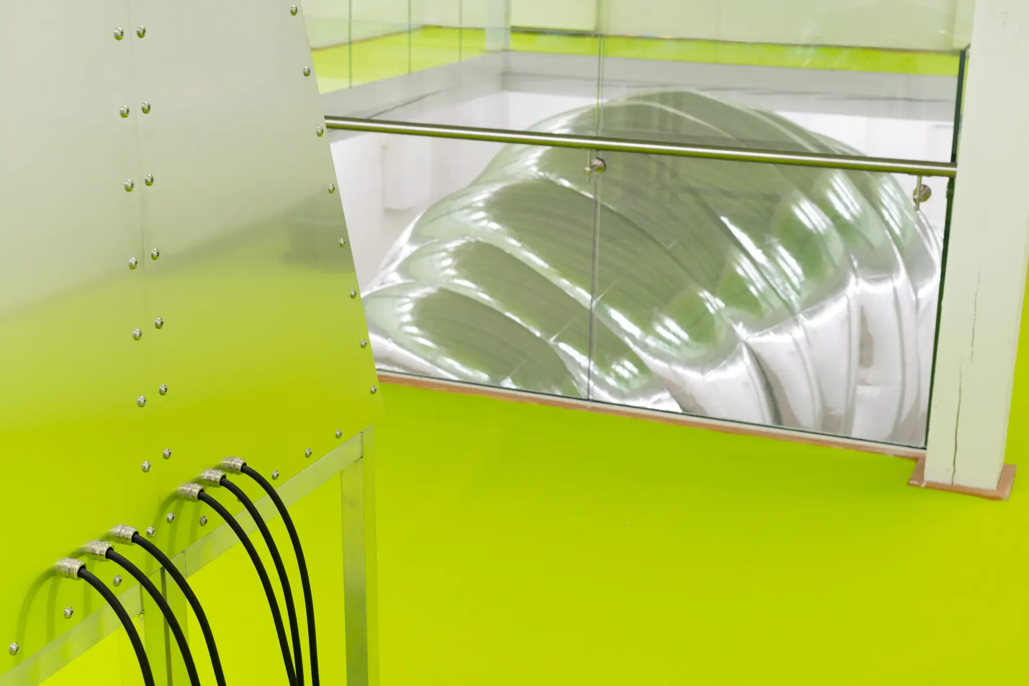 Metal console with connected black cables in foreground and large inflatable silver cocoon sculpture visible below through glass railing, linking interface system and main installation within neon green gallery space, Stine Deja Micro Management Viborg Kunsthal Denmark