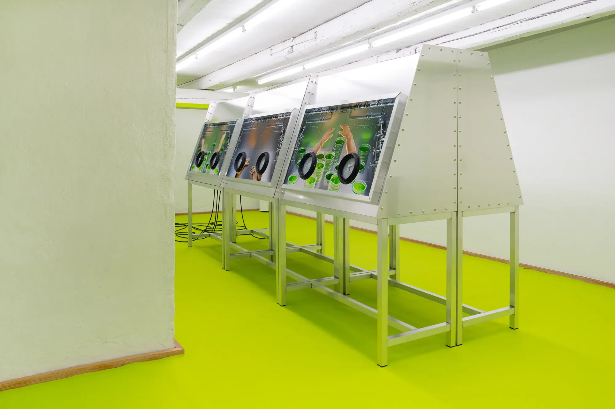 Row of metal console stations with embedded screens showing hands interacting with green liquid elements, installed in bright neon green gallery space with industrial ceiling, Stine Deja Micro Management Viborg Kunsthal Denmark