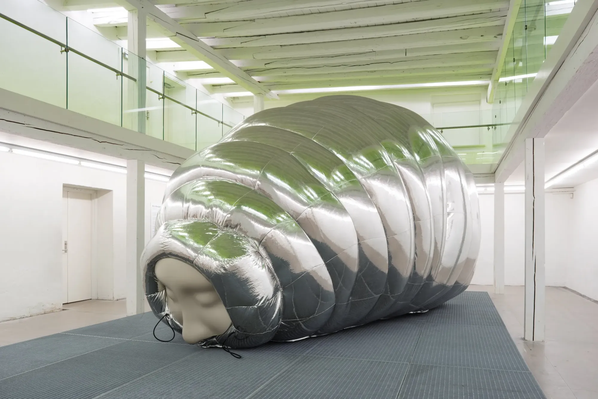 Large inflatable silver cocoon sculpture with human face installed on raised platform within white gallery interior, reflective segmented surface interacting with overhead light and surrounding architecture, Stine Deja Micro Management Viborg Kunsthal Denmark