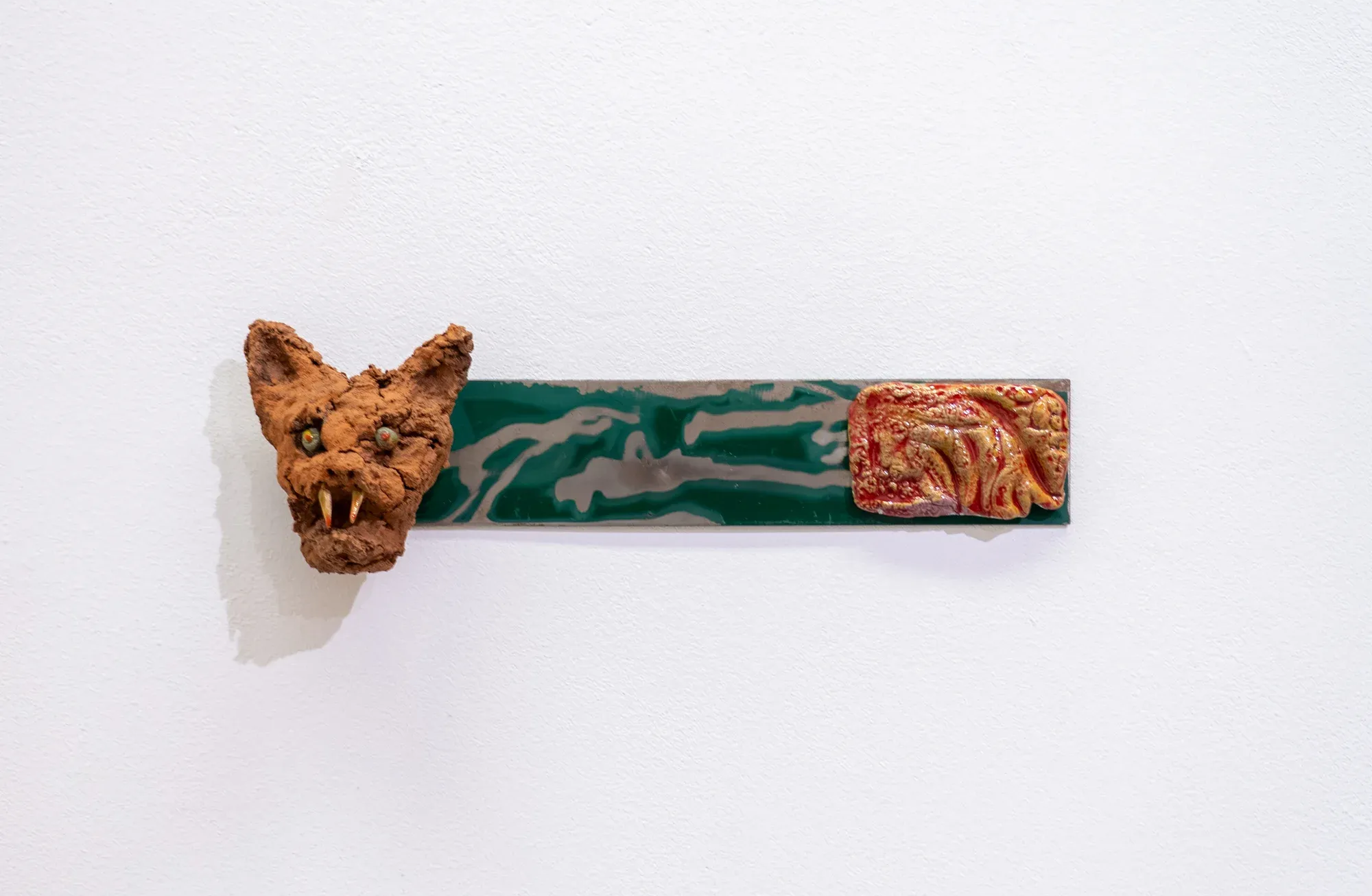 Yorgos Agrotes wall-mounted sculpture shown in Degree Show installation view, small clay bat-like head attached to a green horizontal element on a white wall