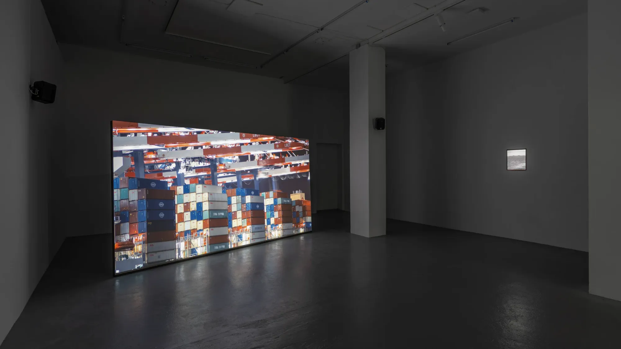 Large illuminated video installation showing stacked shipping containers in dark gallery space, glowing screen casting light across floor, smaller photographic work visible on adjacent wall, Kaya & Blank Intermodal basis Frankfurt 2026 exhibition view