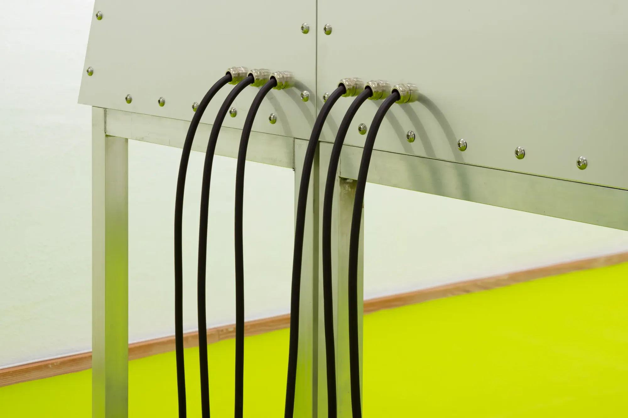 Close-up of industrial metal console with multiple black cables connected via transparent plugs, set against bright neon green floor, detail of interactive installation system, Stine Deja Micro Management Viborg Kunsthal Denmark