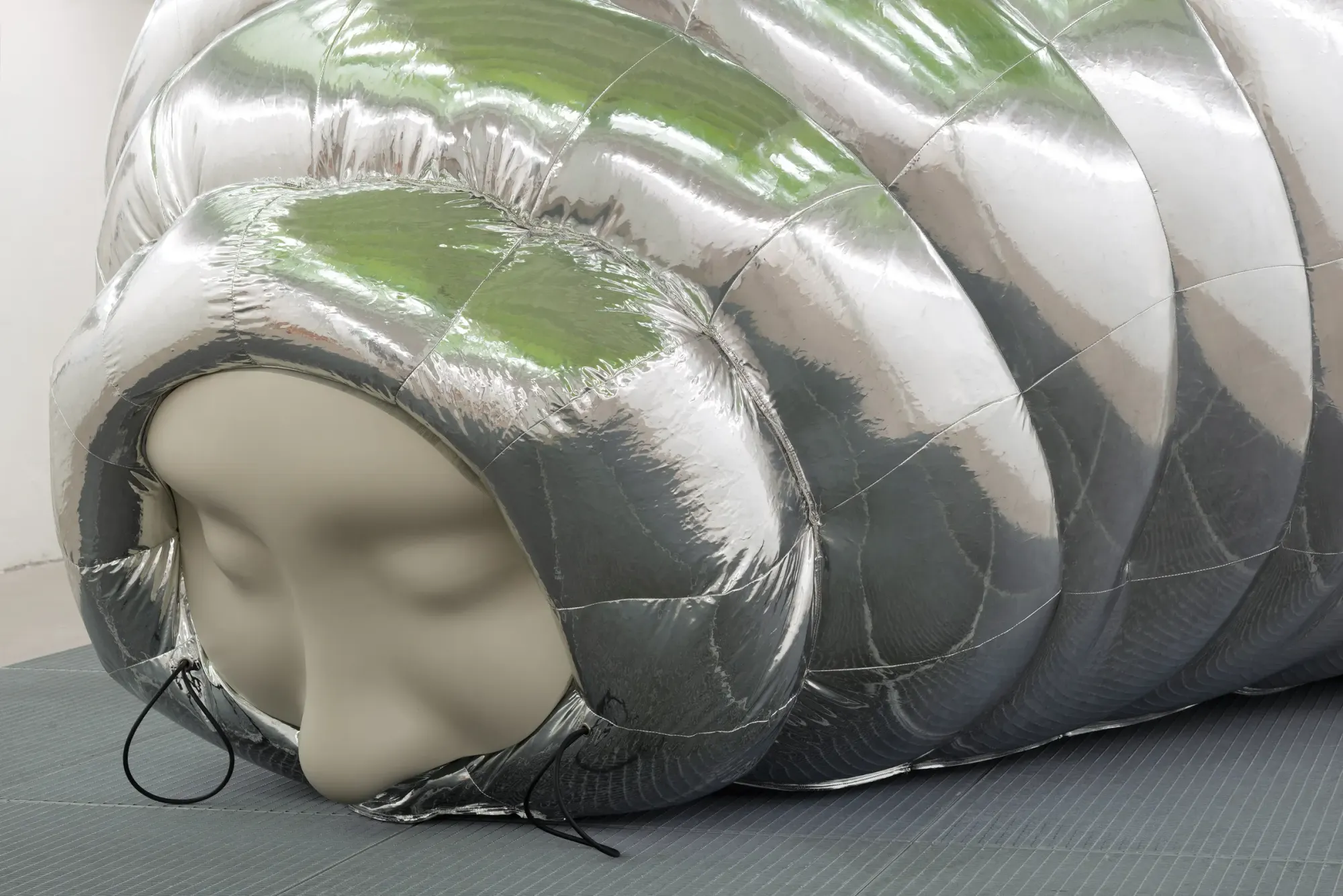 Close-up of inflatable silver cocoon sculpture with smooth human face emerging from segmented reflective surface, resting on dark grid floor, soft matte skin contrasting with metallic texture, Stine Deja Micro Management Viborg Kunsthal Denmar