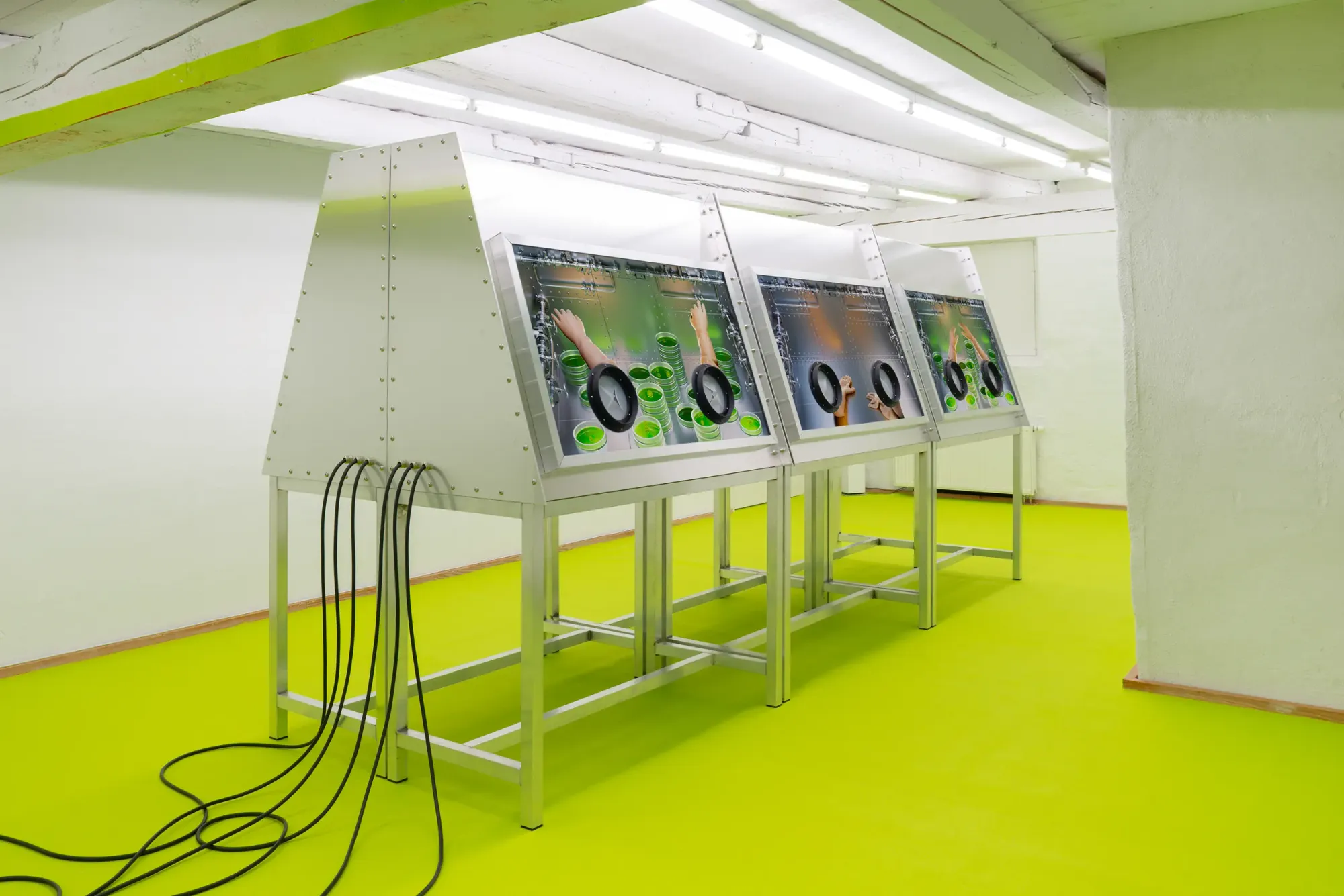 Interactive installation with three metal console stations and embedded screens showing hands interacting with green liquid forms, cables hanging to the floor, set within bright neon green gallery space with white walls and industrial ceiling, Stine Deja Micro Management Viborg Kunsthal Denmark