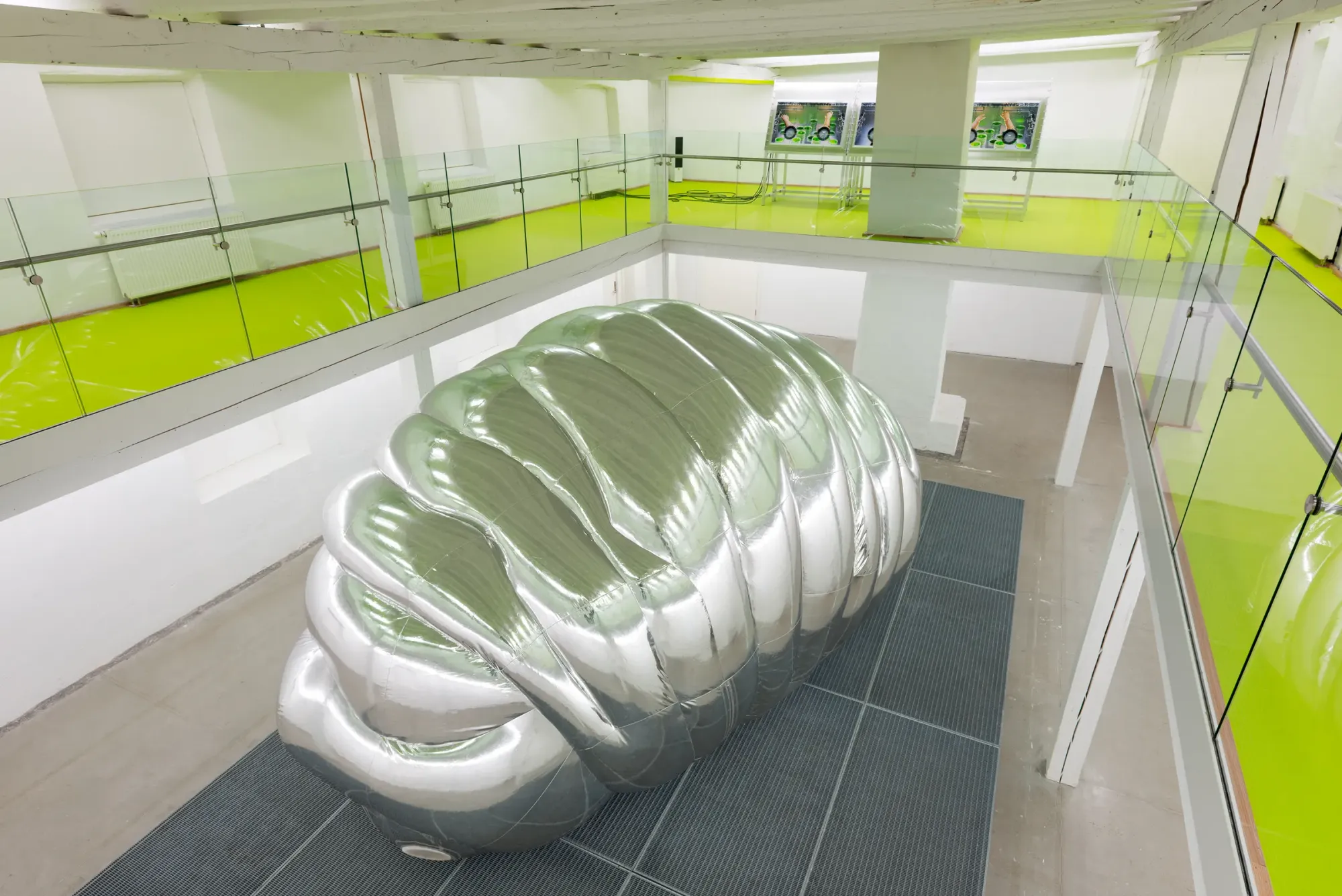 Large-scale inflatable silver cocoon sculpture with segmented metallic surface and hidden human face, installed on dark floor grid inside white gallery space with bright green upper level, Stine Deja Micro Management Viborg Kunsthal Denmark 2026 installation view