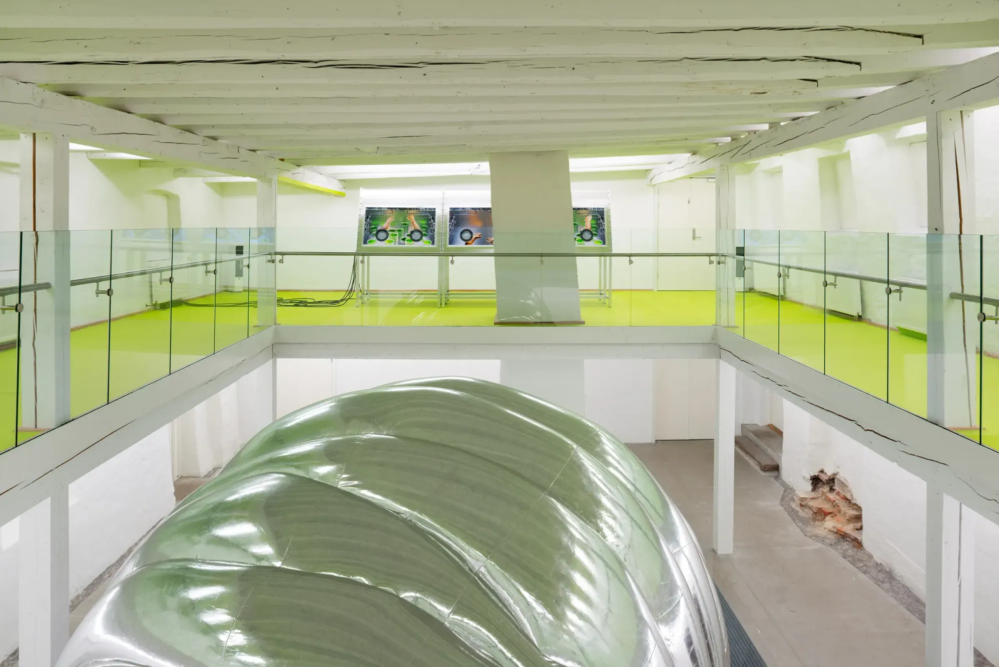 Upper-level view of gallery installation with large inflatable silver cocoon sculpture below and interactive screen consoles in background, bright neon green floor and glass railings defining multi-level exhibition space, Stine Deja Micro Management Viborg Kunsthal Denmark 2026