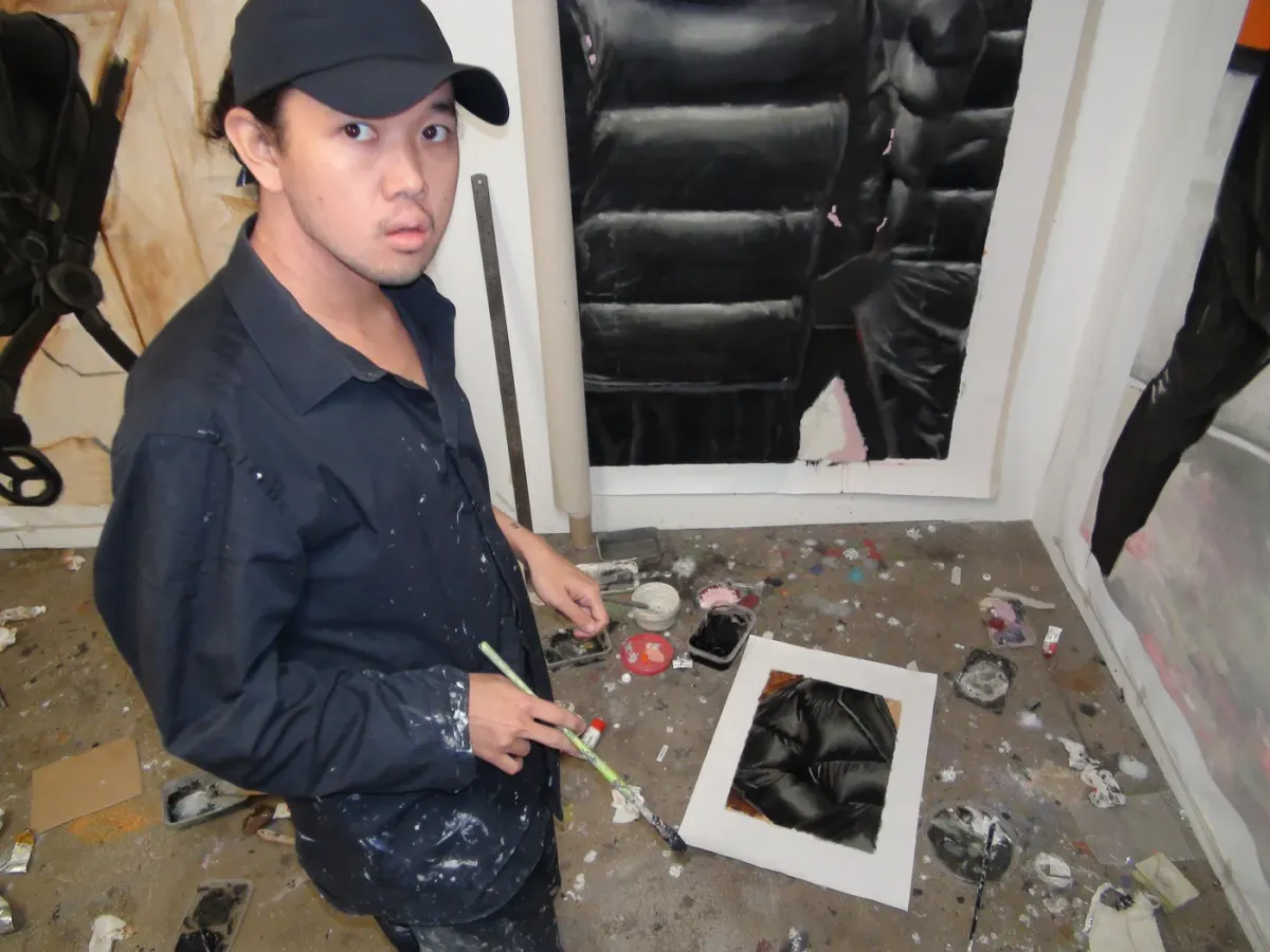 Artist Yutaro Inagaki holding a brush in his studio beside a large puffer jacket painting on the wall.