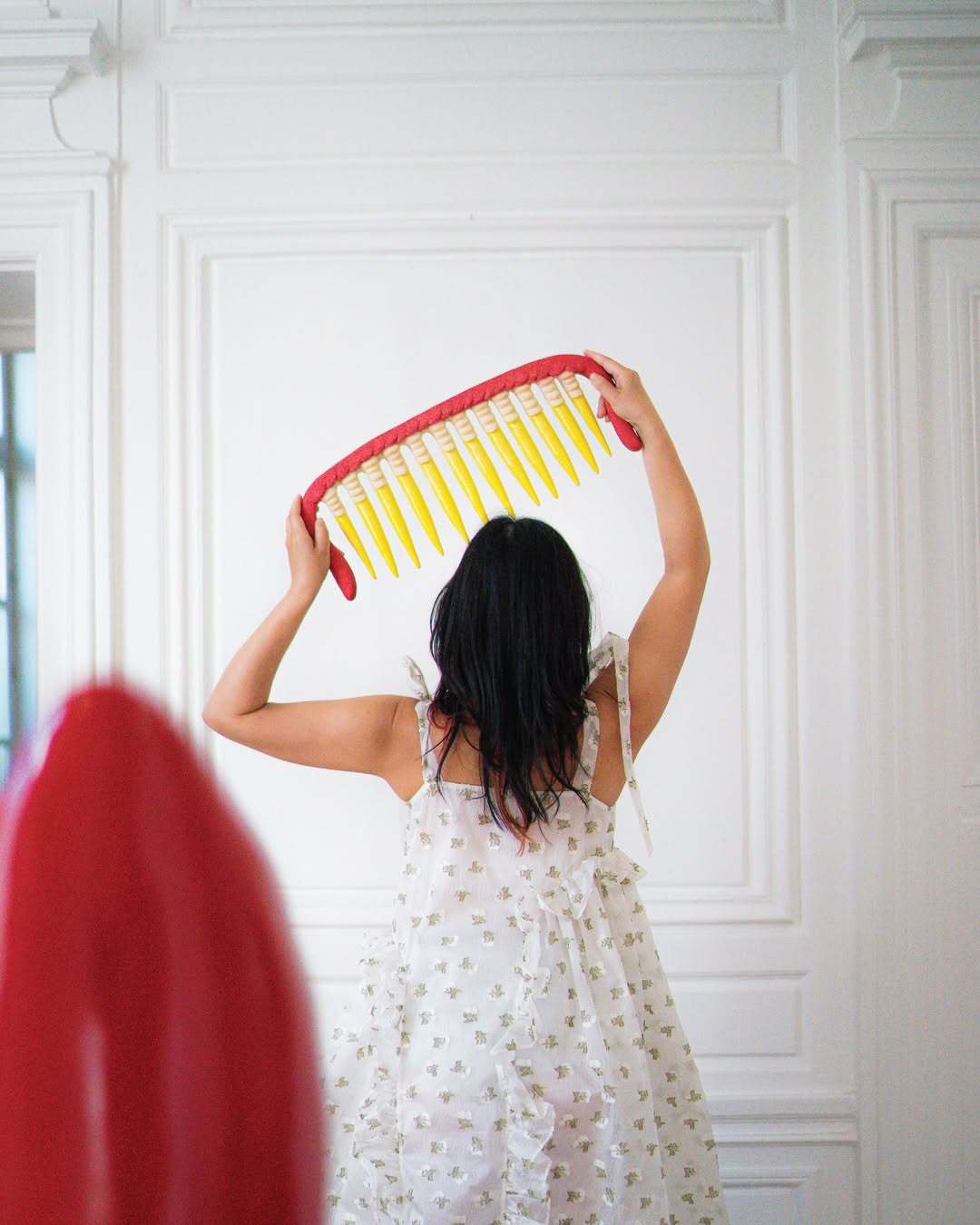 Eri Maeda lifting her large red and yellow ceramic Monster Comb sculpture in a white Paris room, wearing a floral Cecilie Bahnsen dress. Contemporary ceramic art and self presentation.