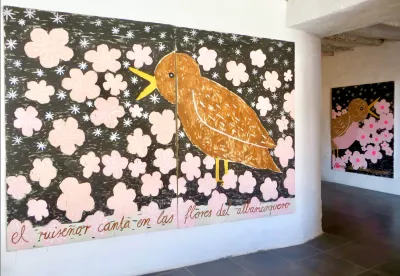 Studio view with two large Gabrielle Grässle canvases: brown bird amid pink blossoms with a handwritten Spanish line; second related painting visible in the adjacent white-walled room.
