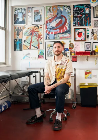 Portrait of artist Billy Bagilhole sitting in his tattoo studio, photographed by Suki Dhanda, surrounded by colourful artworks and drawings on the wall.