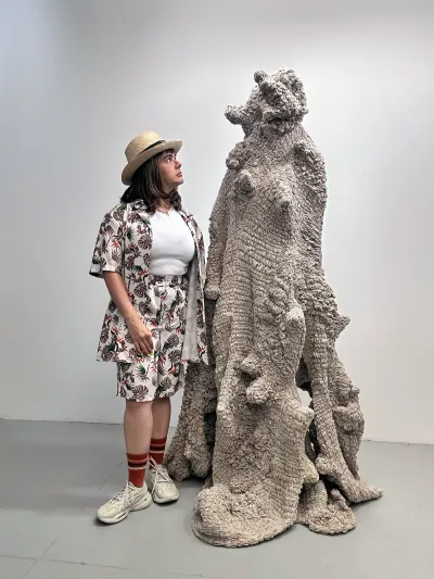 Yali Romagoza standing beside her linen and thread sculpture Landscapes of Belonging: The Body, A Displaced Land (2025), a tall fiber form built from hand-pulled threads and organic textures.
